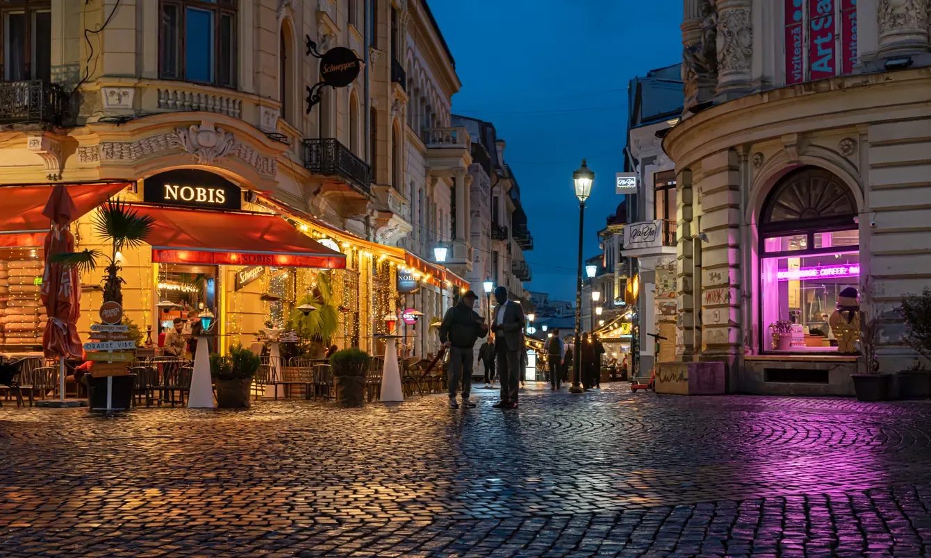 People walking a cobbled street of Bucharest Old Town at blue hour, lit cafe terraces and street lamps, a calm and safe city evening